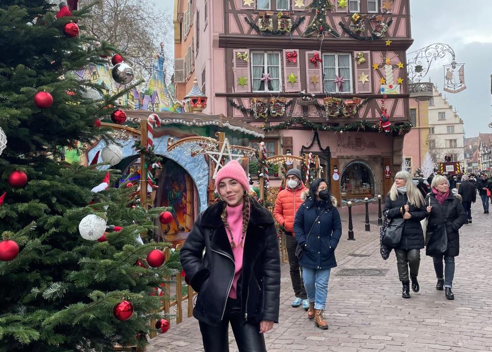 Dasha standing next to a Christmas tree and colorful buildings decorated for Christmas. There are lots of people walking in the area.