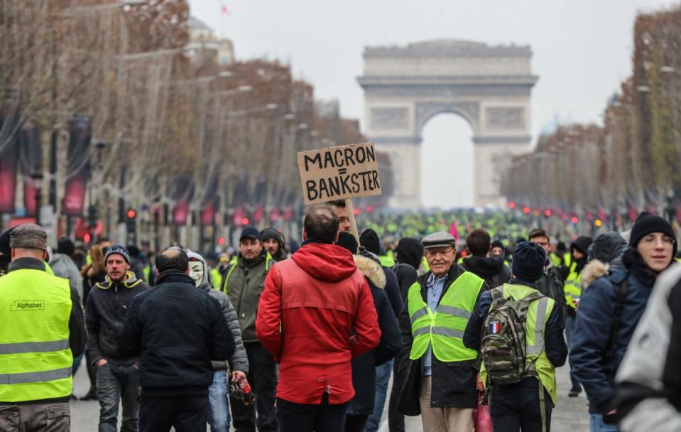 A protester wearing a yellow vest holds a cardboard reading a play on word with