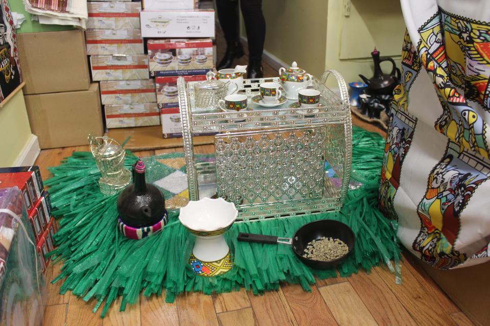 Frehiwot Mamo shows what a traditional Ethiopian coffee ceremony might look like. The green straw mat alludes to the actual grass mat that would be used in Ethiopia. The saucepan in the forefront has pre-roasted coffee beans. The coffee setting in the back shows where people would enjoy the finished product.