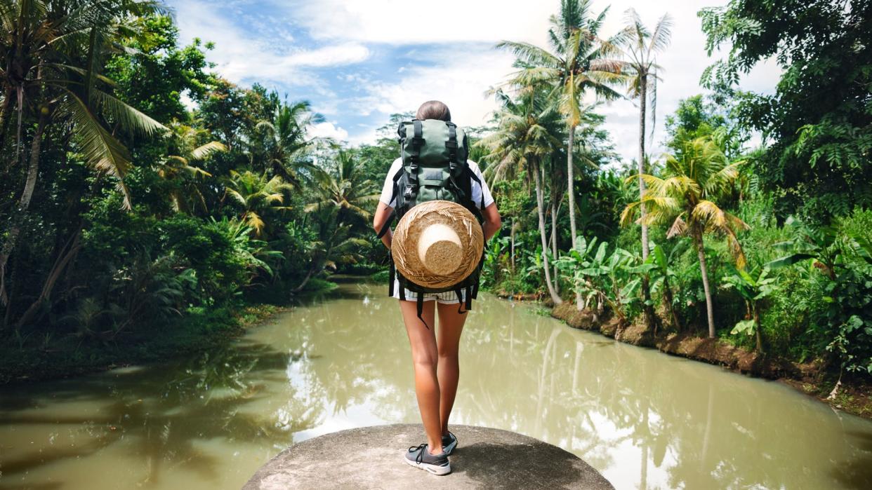 Woman with backpack standing on the edge near big tropical river and looking far away.