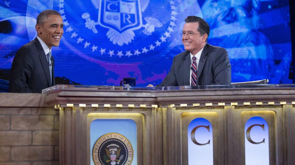 stephen colbert smiling while looking toward barack obama, also smiling, as they sit behind a table for an interview