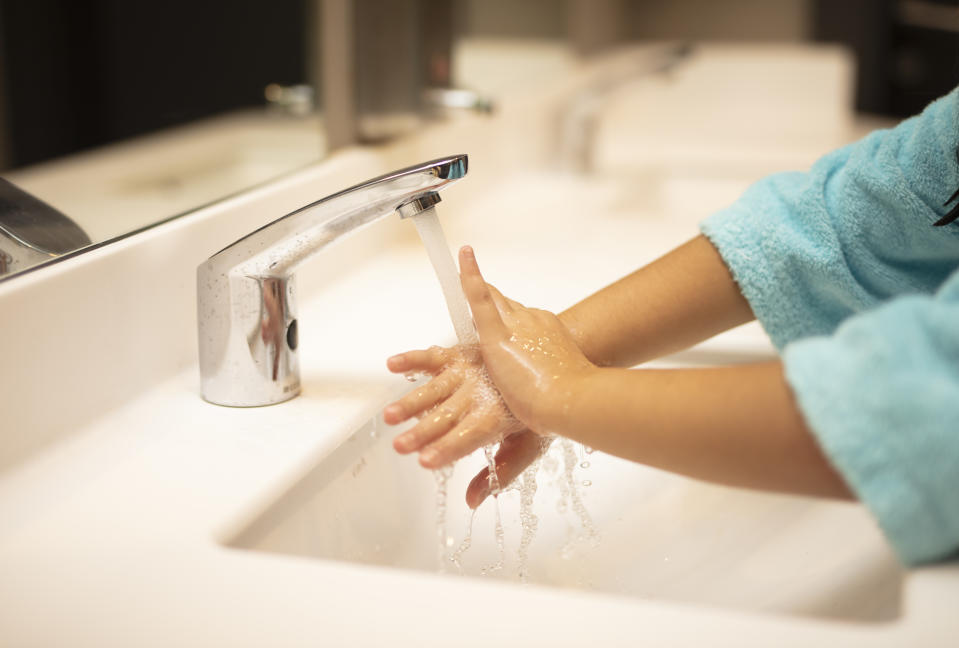 Child washing hands under a faucet in a bathroom sink. Only the child’s hands and part of their arm in a robe are visible