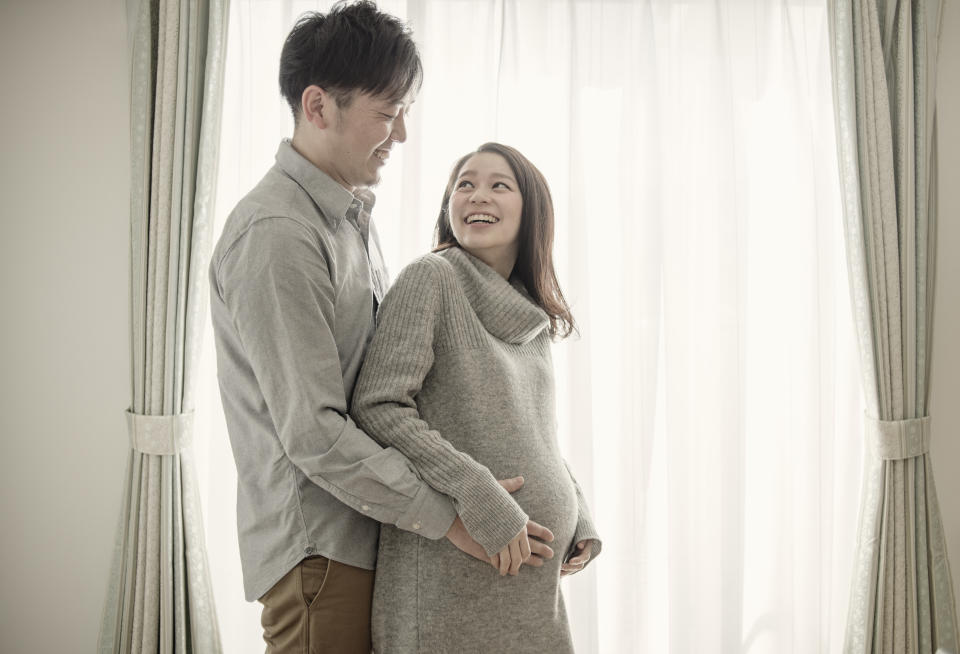 Pregnant young woman standing by window with husband,smiling,holding bump,Japan,Kyoto