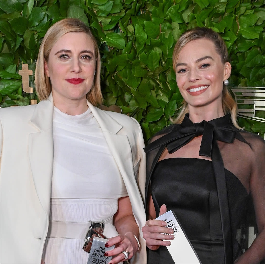  A woman (greta gerwig) in white and a woman (margot robbie) in black, both holding silver awards. 