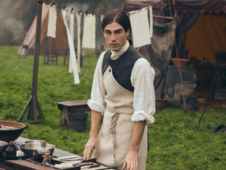 Denzell Hunter standing behind a table with a set of knives and bottles in front of him while wearing a simple outfit with his sleeves rolled up.