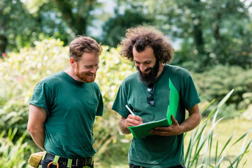 Two men in green shirts go over a document on a clipboard in a garden.