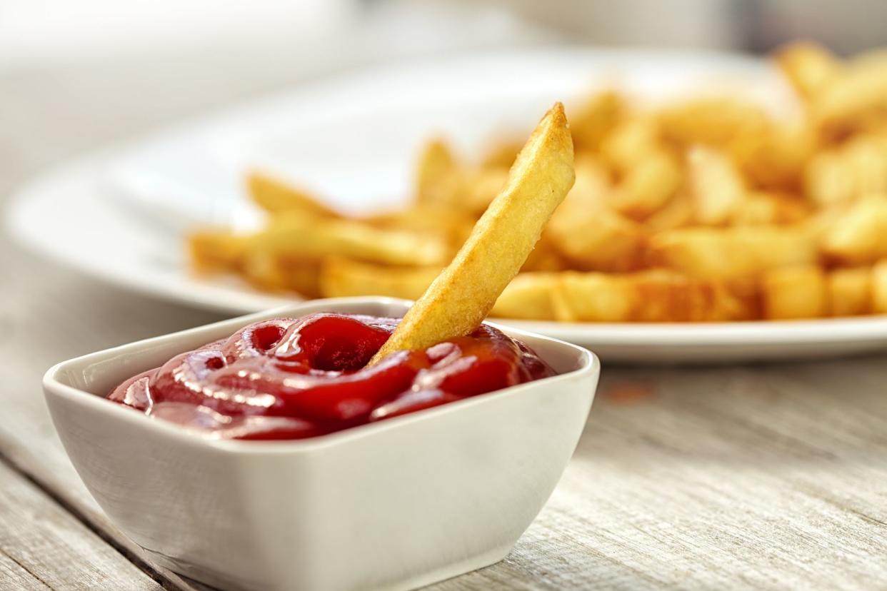 Closeup of one french fry dipped in a square white bowl of ketchup with a blurred backgrounf of a white plate of fries