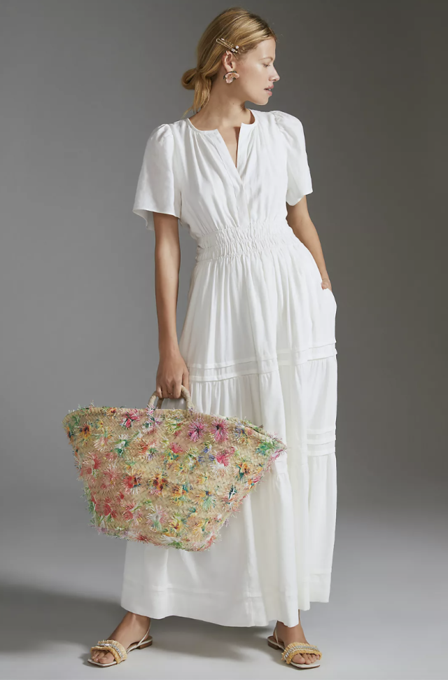 blonde model wearing sandals, carrying straw tote bag and wearing The Somerset Maxi Dress: Linen Edition in white (Photo via Anthropologie)