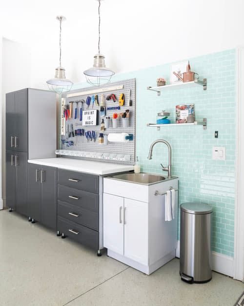 garage with green tiles on the wall, utility sink with a white cabinet under it, next to it is a peg board with a counter top and black cabinets under it