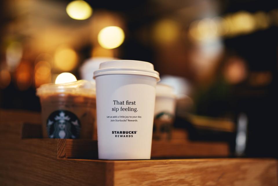 A Starbucks coffee cup, with the Starbucks Rewards logo on it, sitting on a wooden counter.