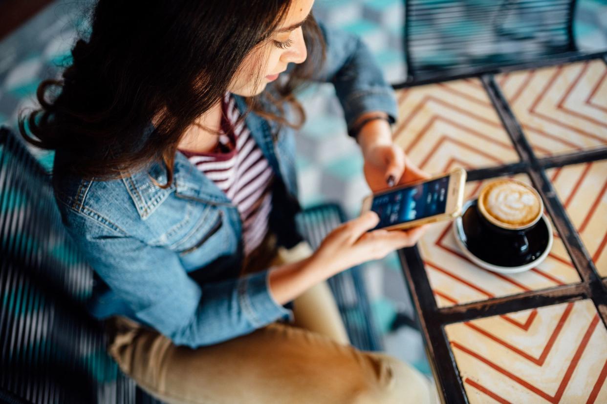 Top view of young woman using a smartphone with both hands while she is sitting in a wire chair at an outdoor cafe, a fancy latte in a black cup and saucer is on a terracotta chevron tile table, blurred floor of patio