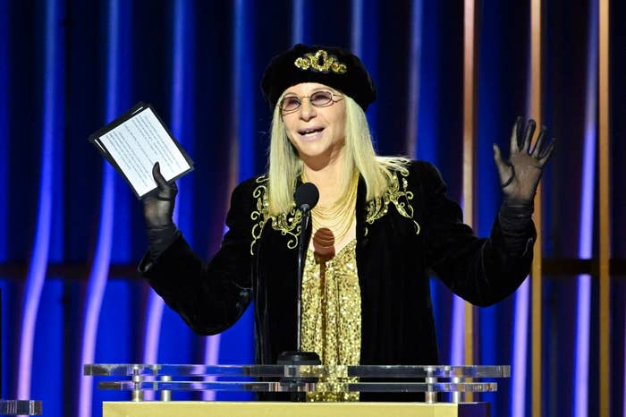 Barbra Streisand on stage at an event, wearing a black jacket with gold embellishments and a hat