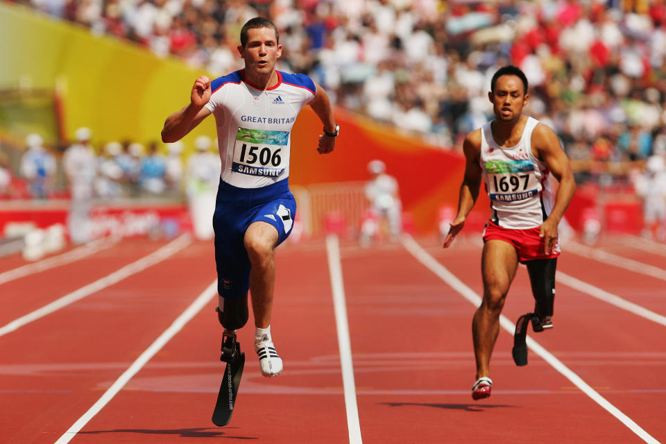 two athletes run at high speed across a constructed track, with a crowd visible far in the back