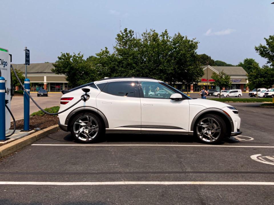 A white Genesis GV60 electric SUV charging at an EVgo station, with trees and sky in the background.