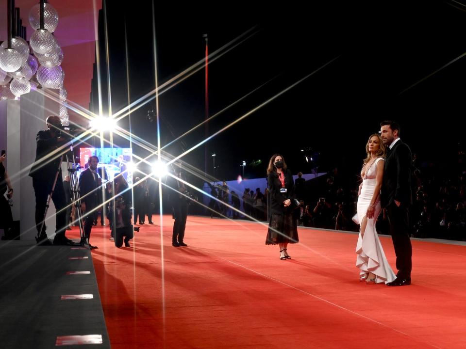 Ben Affleck, wearing a classic black tuxedo, and Jennifer Lopez, wearing a floor-length white mermaid gown, smile as they pose on a red carpet.