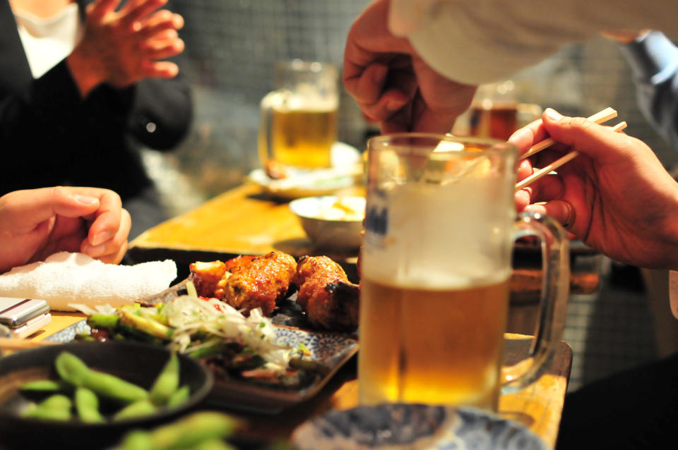 People dining in a restaurant, two hands using chopsticks, plates of food, and glasses of beer on the table. Others are applauding in the background