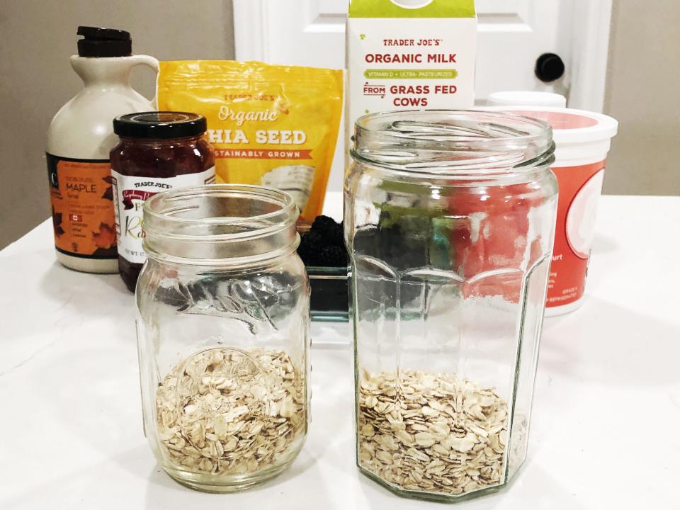 Two jars with oats inside them are placed on a white countertop. Behind them are containers of maple syrup, raspberry preserves, chia seeds, organic milk, Greek yogurt, and fresh berries.