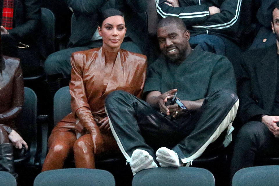 sitting together at a fashion show, Kim, wearing a monochromatic leather suit, appears bored and annoyed while Kanye, wearing a casual tracksuit, puts his feet on the chair in front of him and smiles
