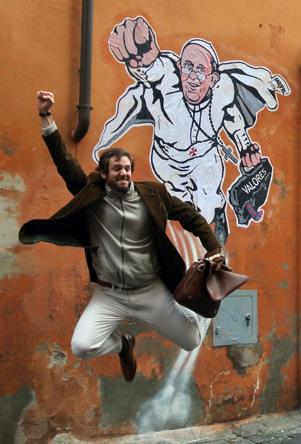 FILE - In this Wednesday, Jan. 29, 2014 file photo, a tourist jumps as poses for a souvenir picture in front of a graffiti by Italian artist Mauro Pallotta depicting Pope Francis as Superman and holding a bag with a writing which reads: "Values" at the Borgo Pio district near St. Peter's Square in Rome. Pope Francis’ status as a superhero has bit the dust. Rome’s decorum police early Thursday, Jan. 30, 2014, scrubbed the wall near the Vatican where “SuperPope” had been displayed, showing Francis as Superman in flight and clutching his black satchel of values to spread to the world. Artist Mauro Pallotta had put the image up on Monday in homage to Francis. Pallotta’s agent, Margaret Porpiglia said Thursday the artist is now hoping to avoid a city fine but is considering making a street art piece depicting the “anti-hero” Rome Mayor Ignazio Marino. The white caped crusader pope, which was actually painted on paper and affixed to the wall with water-based glue, had drawn immense popular interest around the Borgo neighborhood of tiny cobble stoned streets near St. Peter’s Square. (AP Photo/Gregorio Borgia, File)