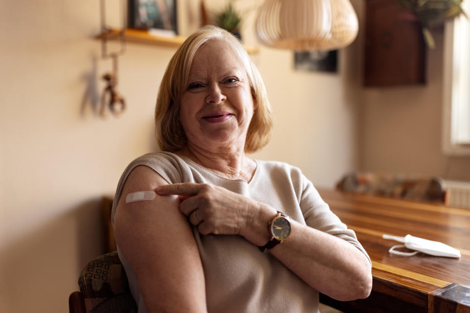 Senior female looking at camera pointing at her arm with adhesive bandage after getting the covid-19 vaccine. Mature woman feeling happy after getting coronavirus vaccine at home.