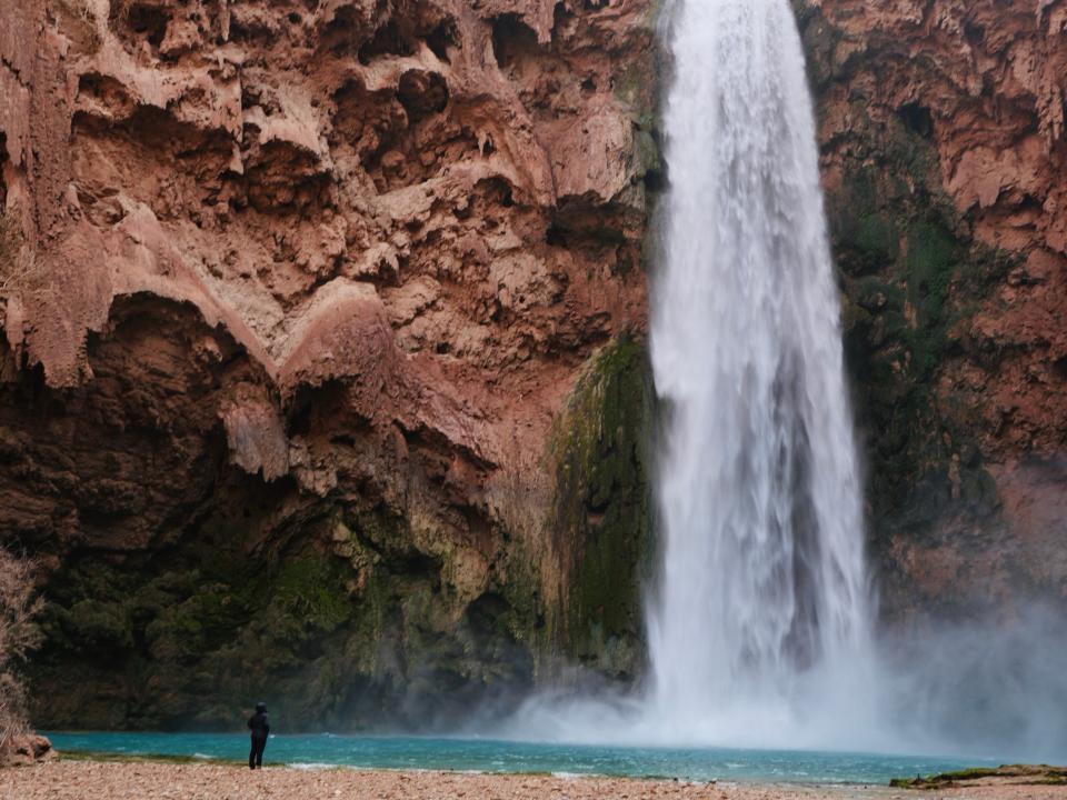 A person looks at Mooney Falls, a massive waterfall cascading down red rocks into blue-green water.