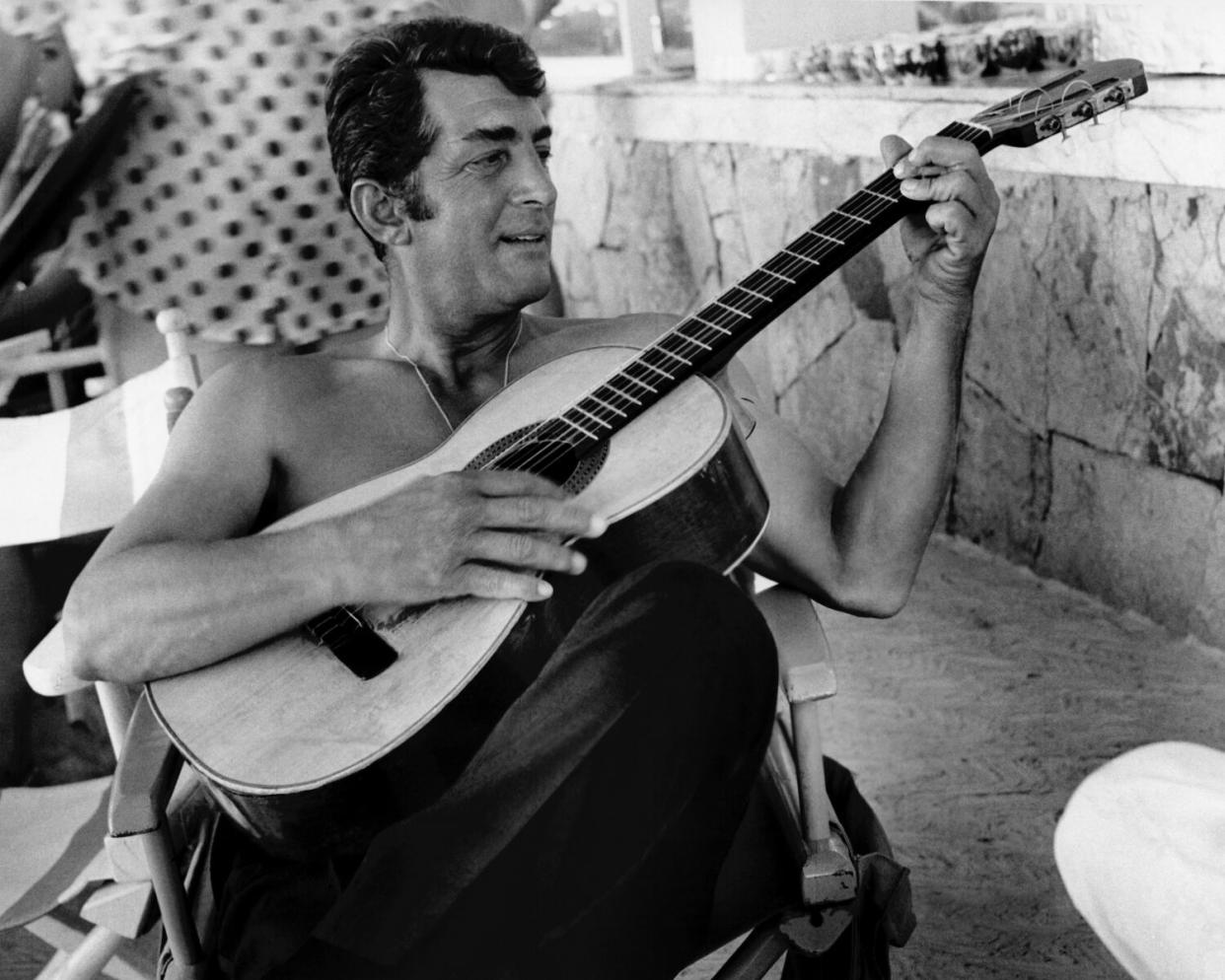 A man, shirtless, seated and playing the acoustic guitar