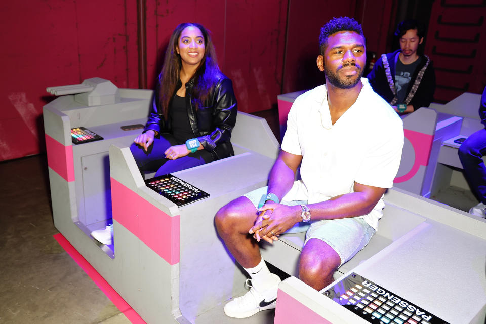 Two people seated in futuristic chairs, man in shorts and a woman in a leather jacket, with interactive displays in front