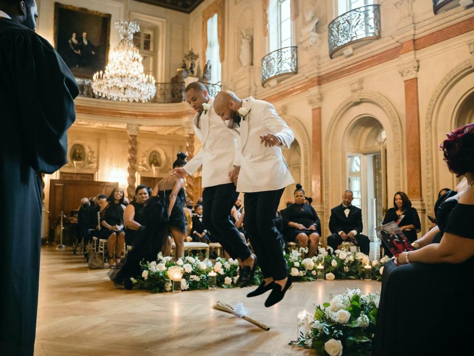 Two grooms jump the broom at their wedding wearing white tuxedos.