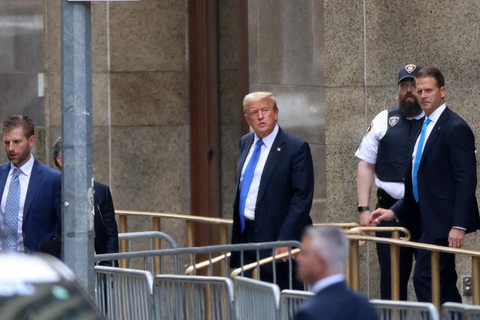 Donald Trump walks near a building entrance, surrounded by security personnel and officials in suits