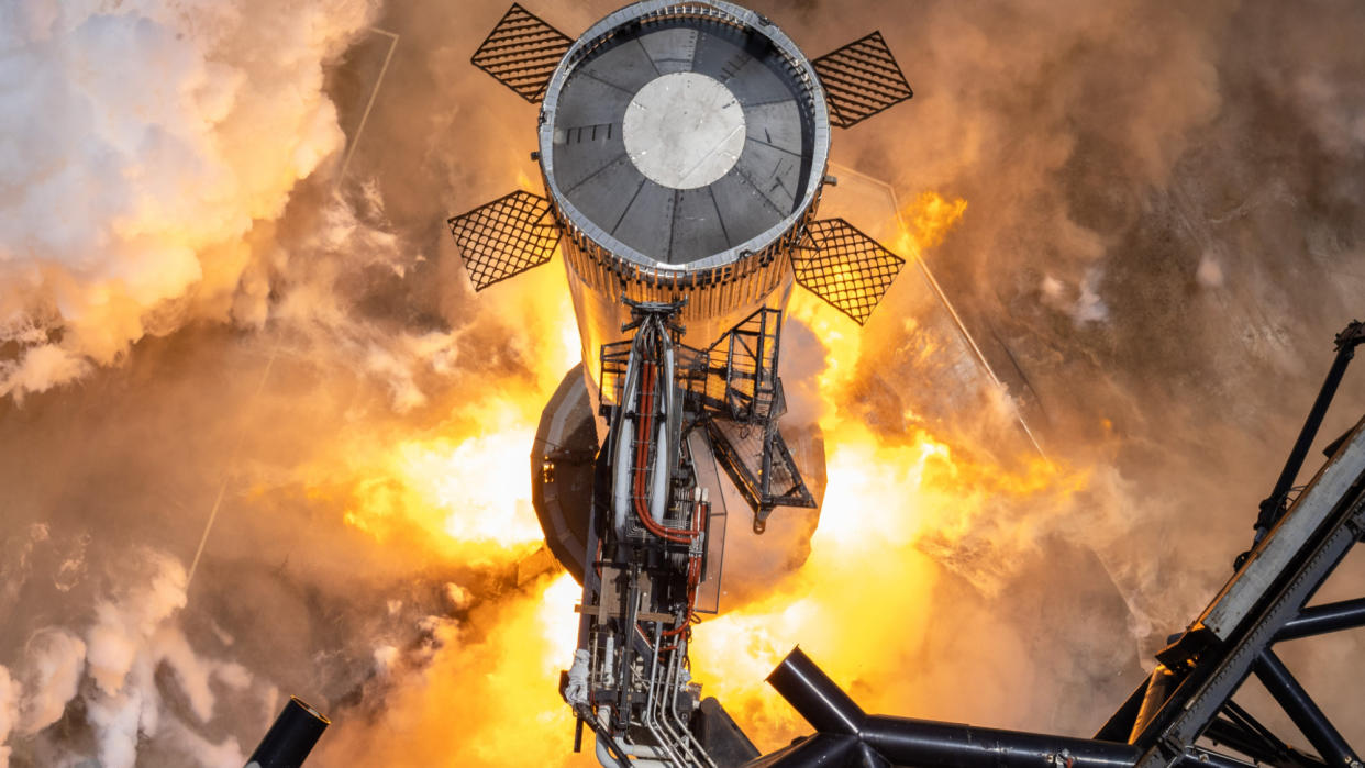  flames erupt from the base of a shiny silver rocket, as seen from above, during an engine test. 