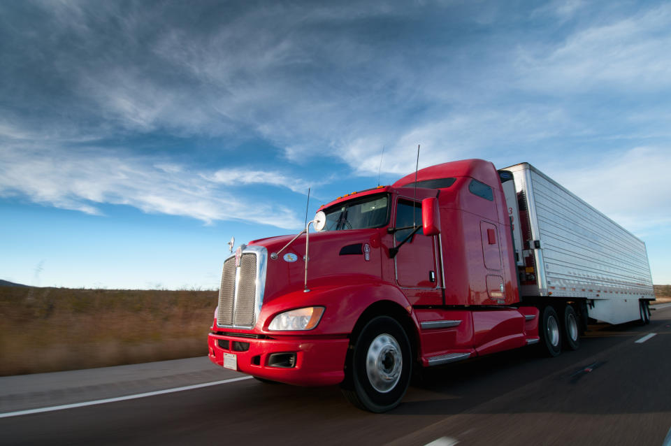 A red semi-truck drives on a highway under a cloudy sky, with a focus on the truck's design and the open road. There are no people in the image