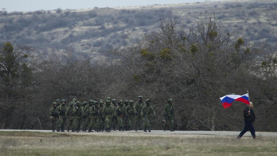 Russian solders walk as a local resident waves with Russian flag outside of a Ukrainian military base in the village of Perevalne, outside of Simferopol, Ukraine, on Sunday, March 2, 2014. Hundreds of armed men in trucks and armored vehicles surrounded the Ukrainian military base Sunday in Crimea, blocking its soldiers from leaving. (AP Photo/Ivan Sekretarev)