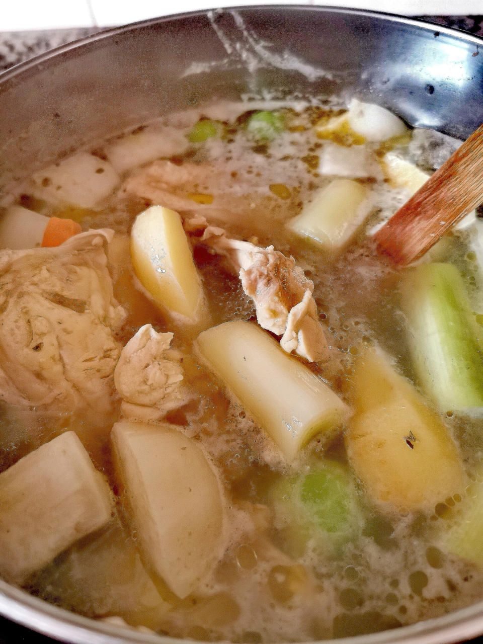 A pot of soup with large chunks of vegetables, such as potatoes and celery, and pieces of chicken being stirred by a wooden spoon