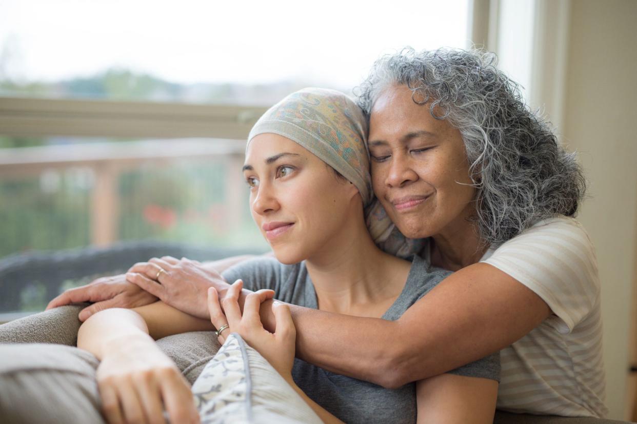 Hawaiian woman in 50s embracing her mid-20s daughter on couch who is fighting cancer