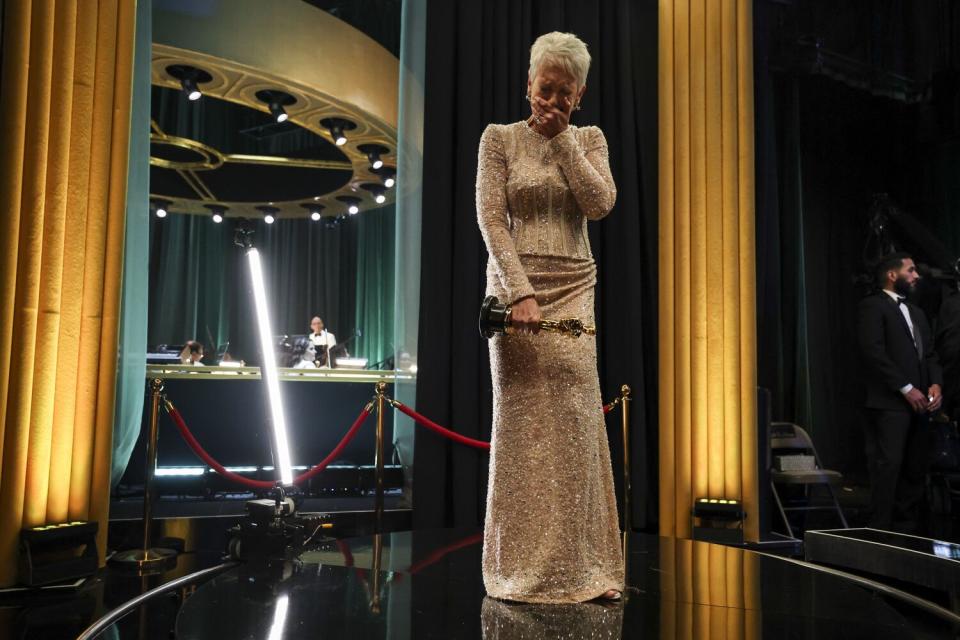 A woman in a champagne colored dress holds an Oscar and cries backstage.