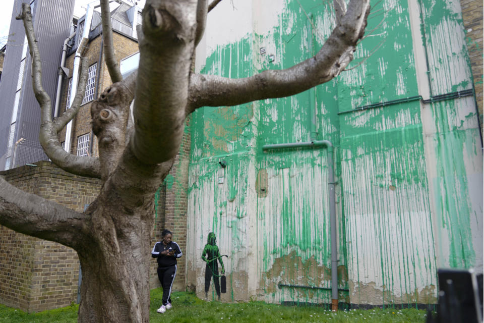 A woman looks at a new Banksy painting on a wall in London, Monday, March 18, 2024. A new Banksy mural drew crowds to a London street on Monday, even before the elusive graffiti artist confirmed that the work was his. The artwork in the Finsbury Park neighborhood covers the wall of a four-story building and shows a small figure holding a pressure hose beside a real tree. Green paint has been sprayed across the wall, replicating the absent leaves of the tree, which has been severely cropped. Banksy claimed the work by posting before and after photos of the location on his official Instagram account.(AP Photo/Alastair Grant)