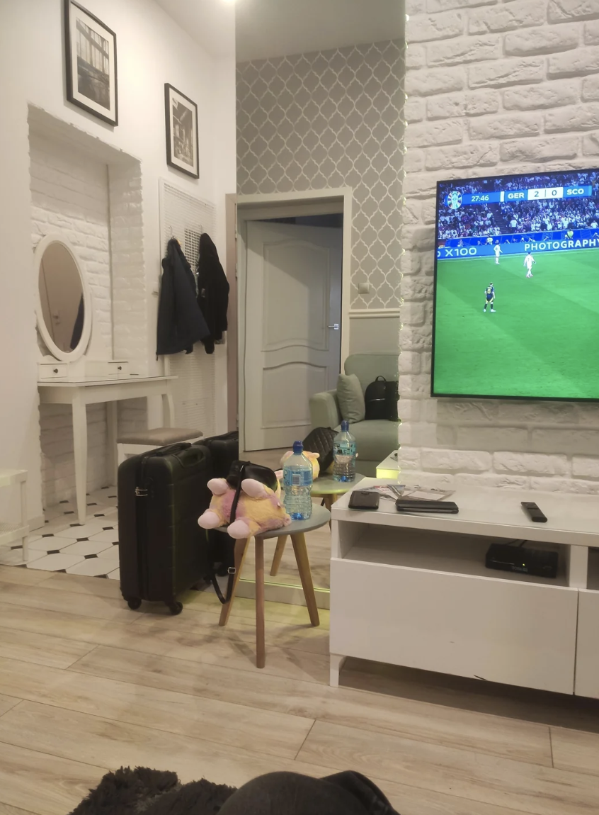 A living room with a television showing a soccer game, a mirror on the wall, suitcases, pink slippers on a table, and water bottles on a table by the door