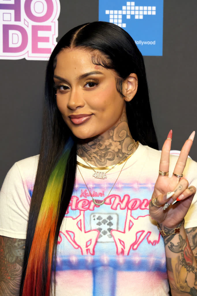 Kehlani poses on the red carpet wearing a graphic t-shirt, displaying long, straight hair with rainbow accents. She is holding up a peace sign