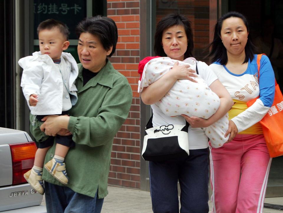 A woman holds a young boy, left, and another holds a swaddled infant next to a young woman outside a building