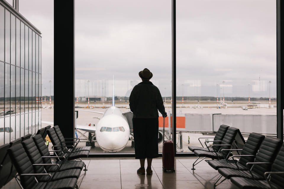 Silhouette of a person wearing a hat with hand luggage in international airport, looking through the window at planes, travel concept