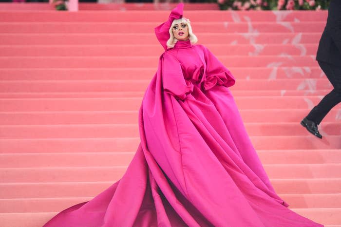 Lady Gaga poses on stairs in a voluminous, flowing gown with a high neckline and oversized sleeves, accessorized with a matching headpiece