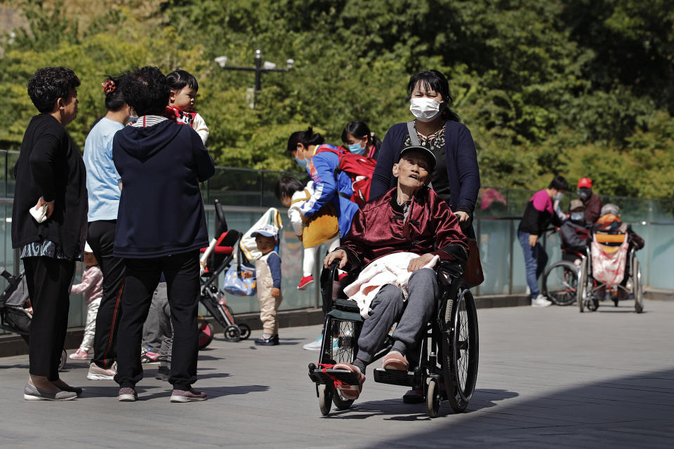 A woman wearing a face mask pushes an elderly man on a wheelchair passes by residents play with their children near a commercial office building in Beijing on Monday, May 10, 2021. China’s population growth is falling closer to zero as fewer couples have children, the government announced Tuesday, adding to strains on an aging society with a shrinking workforce. (AP Photo/Andy Wong)