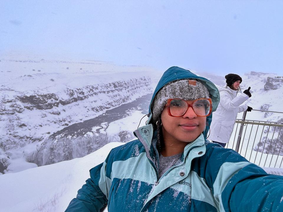The writer wears a blue coat, gray hat, and red-frame glasses and stands on a snowy mountain in Iceland