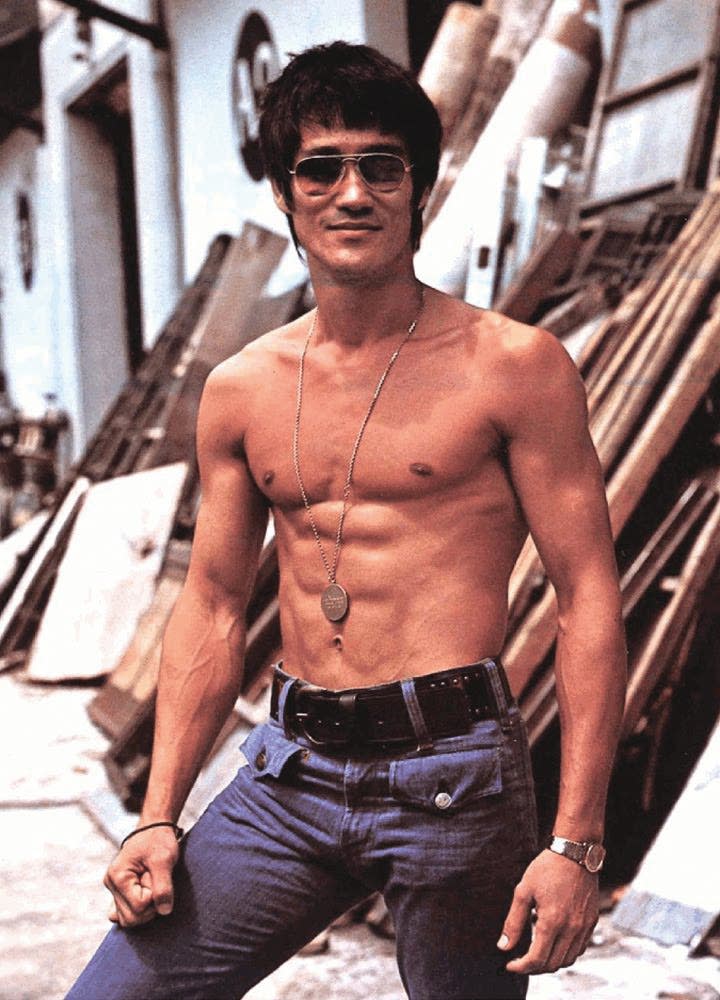 Bruce shirtless, wearing medallion necklace, sunglasses, smiling, standing before set construction.