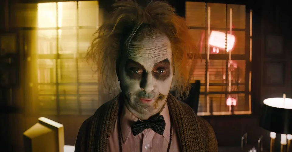 Michael Keaton as Beetlejuice, with disheveled hair and worn-out makeup, in a dimly lit room with a backdrop of bright lighting through window blinds