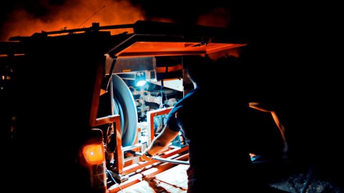 Man operating equipment nearby to fire. It is dark, but you can see an orange glimmer of the fire in the background.