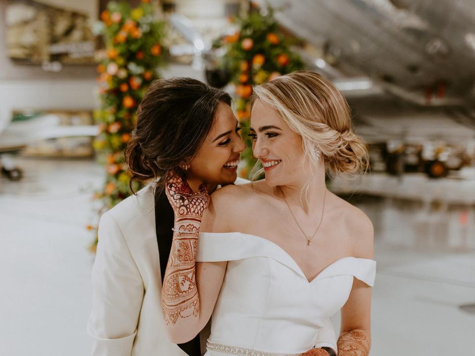 A bride hugs her wife from behind as her wife cups her face.