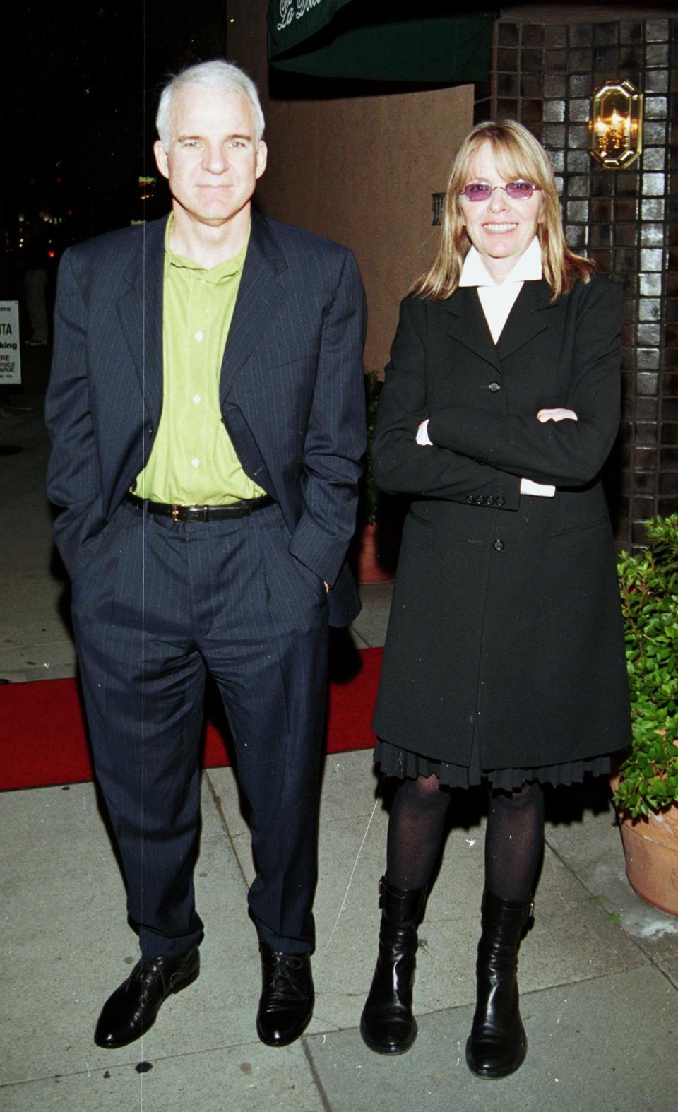 Steve Martin and a female companion stand together; he wears a suit and she's in a coat and knee-high boots