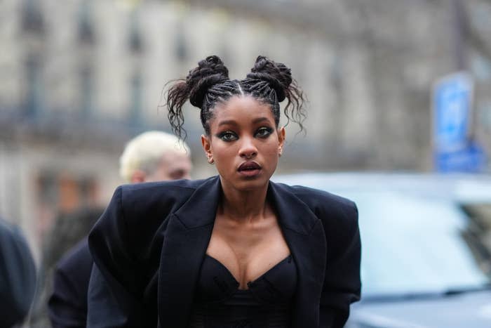 Person in a stylish black suit with unique bun hairstyle walking outdoors