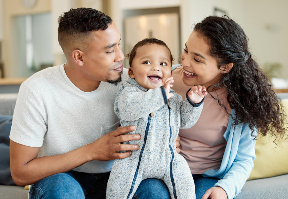 A smiling couple sits on a couch, holding a happy baby between them. All are casually dressed. Names not known